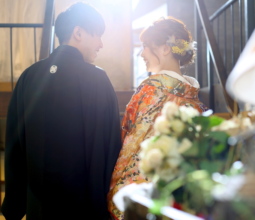 花々を手前に、寄り添って歩く後ろ姿の新郎新婦、黒紋付の新郎と鮮やかなオレンジと金の色打掛姿の新婦、スタジオでの撮影。