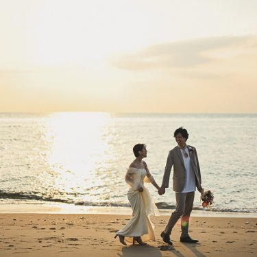 夕日が映る海をバックに手をつないで砂浜を歩く新郎新婦