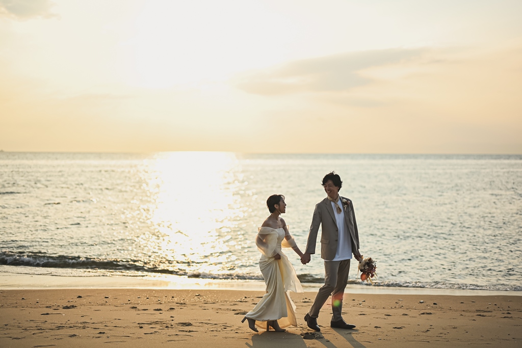 夕日が映る海をバックに手をつないで砂浜を歩く新郎新婦