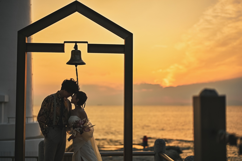 夕焼け空バックに鐘の前でおでこを合わせる新郎新婦