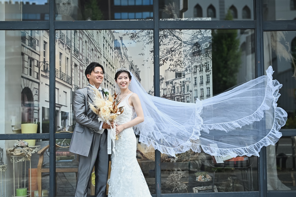 外観バックにヴェールをなびかせ景色を眺める新郎新婦