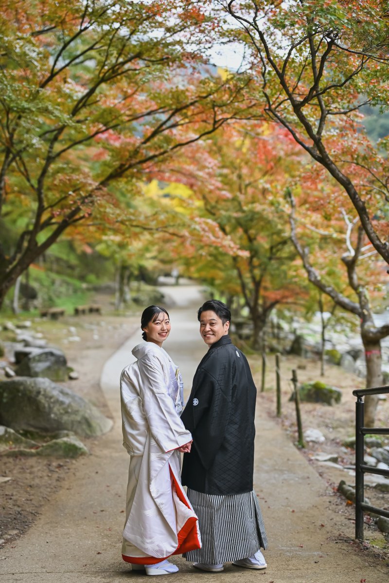 紅葉のトンネルの下を手を取り合って歩くポーズの新郎新婦