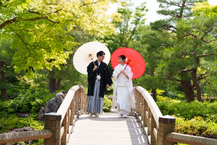 池に架かる橋の上で赤と白の和傘を差し手をつなぐ白無垢の新婦と紋付袴の新郎、緑豊かな庭園での和装前撮り