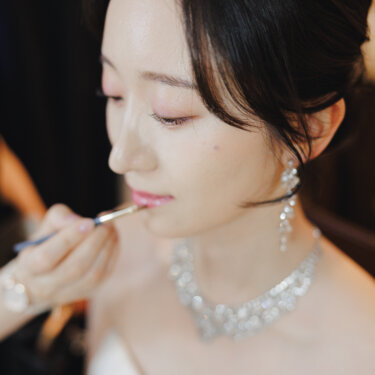 ティアラとネックレスをつけた花嫁がリップを塗ってもらう、ヘアメイクの支度シーン。フォトウェディング当日の一枚
