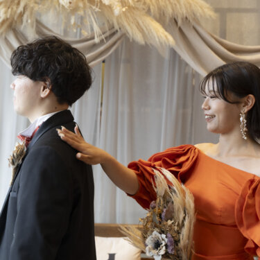 背を向けた新郎の肩をそっとたたく、オレンジのパフスリーブのカラードレスの花嫁。ファーストミートのウェディングフォト