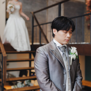 目を閉じて待つグレーのフロックコートの新郎と、階段を下りてくるウェディングドレスの花嫁。ファーストミートの前撮り