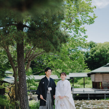 松の木と赤い縁台のある庭園で、池を背に並んで立つ紋付袴の新郎と白無垢の花嫁を引きで写した和装ロケーションフォト