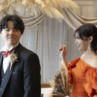 背を向けて待つ赤い蝶ネクタイの新郎と、後ろで手を上げるオレンジのカラードレスの花嫁。ファーストミート直前の前撮り
