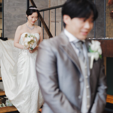 手前の新郎をぼかし、トレーンを広げて階段を下りてくるティアラの花嫁を写したファーストミートのウェディングフォト