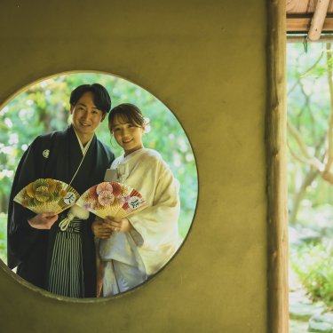 白鳥庭園の円窓から、扇子を持って寄り添う新郎新婦をのぞき見るように写した和装フォトウェディング