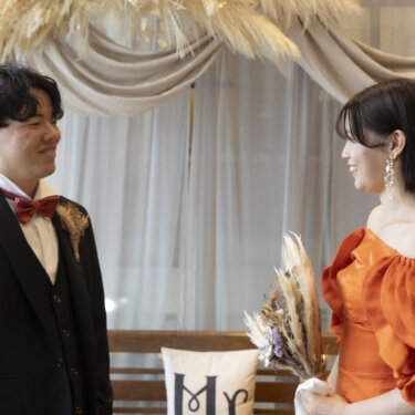 パンパスグラスのドレープの前で、振り返った新郎とオレンジのカラードレスの花嫁が見つめ合うファーストミートのスタジオフォト