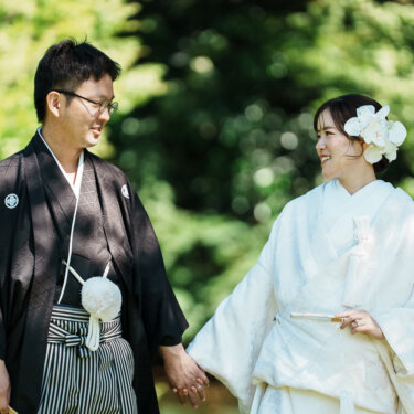 木漏れ日の中で手をつなぎ、見つめ合って笑う紋付袴の新郎と白無垢の花嫁の和装前撮り