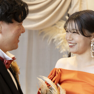 パンパスグラスのドレープの前で見つめ合って笑う、赤い蝶ネクタイの新郎とオレンジのカラードレスの花嫁の前撮り