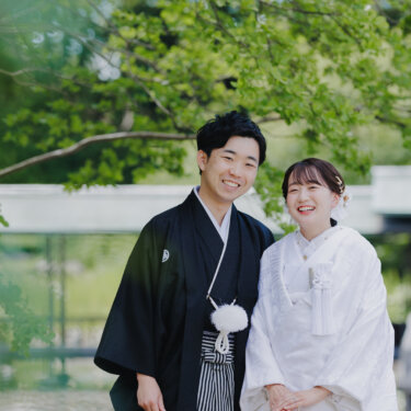 池と屋根付きの廊下を背に、並んでカメラに笑う紋付袴の新郎と白無垢の花嫁の和装前撮り