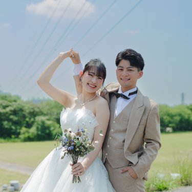 青空の下、ウェディングドレスの花嫁と新郎が腕を合わせてハートを作り、カメラに笑顔を向けるウェディングフォト