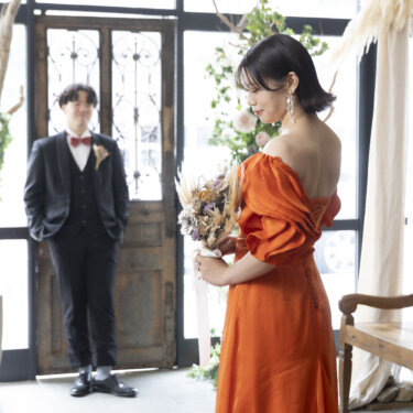 アンティークの扉の前に立つ新郎と、背中の編み上げを見せるオレンジのカラードレスの花嫁。ファーストミートのスタジオフォト