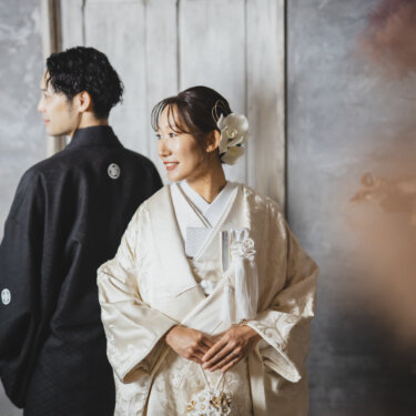 グレーの壁の前で、奥を見る新郎と花の髪飾りをつけて横を向く白無垢の花嫁。落ち着いた雰囲気の和装前撮り