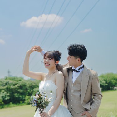 青空の河川敷で、腕でハートの形を作ってほほえむ花嫁と新郎。人気のハートポーズの前撮り