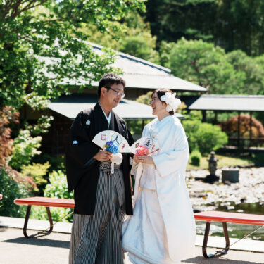 赤い縁台の並ぶ庭園で、'夫''妻'の扇子を持って笑い合う紋付袴の新郎と白無垢の花嫁の和装フォトウェディング