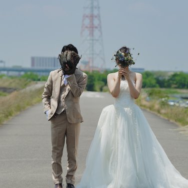 鉄塔の立つ河川敷の道で、グローブとブーケで顔を隠す新郎新婦。遊び心ある顔隠しポーズの前撮り