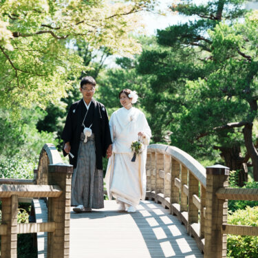 緑に囲まれた木の橋を並んで渡る新郎新婦を正面から写した和装前撮り。夏の日本庭園の一枚