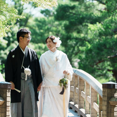 木の反り橋を並んで歩き、笑い合う紋付袴の新郎とブーケを持つ白無垢の花嫁。白鳥庭園の和装フォトウェディング