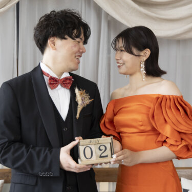 'JULY 02'の日付ブロックを二人で持ち、見つめ合う赤い蝶ネクタイの新郎とオレンジのカラードレスの花嫁の前撮り