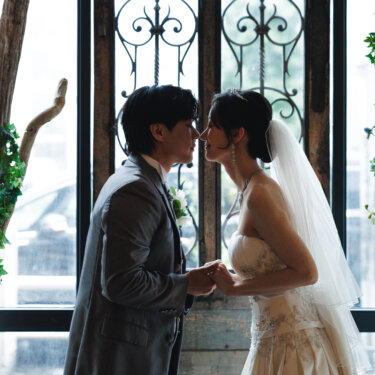 アイアン装飾の窓と花のアーチの前で、ベールをつけた花嫁と新郎がキスをする逆光のウェディングフォト