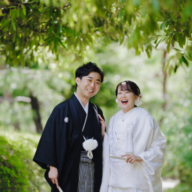 新緑の木々の下で、扇子を持って笑い合う紋付袴の新郎と白無垢の花嫁の和装フォトウェディング