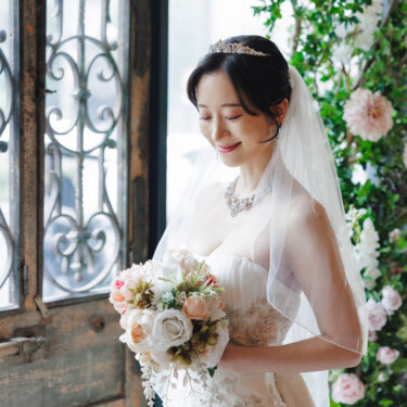 アイアン装飾の窓と花のアーチの前で、ブーケを見つめてほほえむベールとティアラの花嫁のウェディングフォト