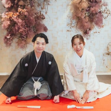 ピンクのドライフラワーの前の赤い毛氈に並んで正座し、カメラに笑う新郎新婦の和装前撮り