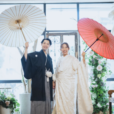 白と赤の和傘を高く掲げて笑う紋付袴の新郎と白無垢の花嫁。シャンデリアのあるスタジオの和装フォトウェディング