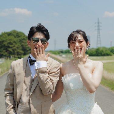 青空の河川敷で口元を手で隠して笑う、サングラスの新郎新婦の正面カット。遊び心あるウェディングフォト