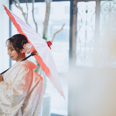 赤い和傘を肩にかけ、振り返ってほほえむ白無垢の花嫁。窓辺の光がやわらかな和装前撮り