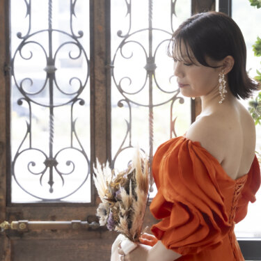 アイアンの窓の前で、背中の編み上げを見せてうつむくオレンジのカラードレスの花嫁。バックスタイルが映える前撮り