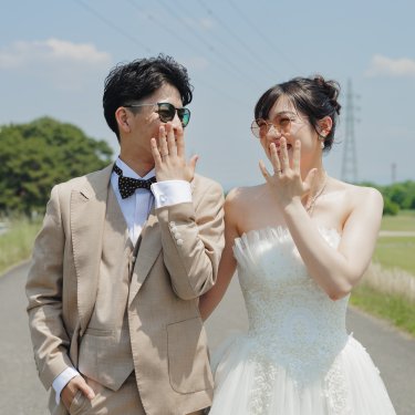 河川敷の道で口元を手で隠し、照れ笑いする新郎とサングラスの花嫁。青空が広がる前撮り