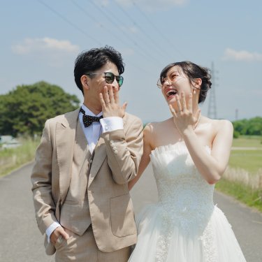 青空の下の河川敷の道で、口元に手を当てて笑い合うサングラスの新郎新婦。自然体のウェディングフォト