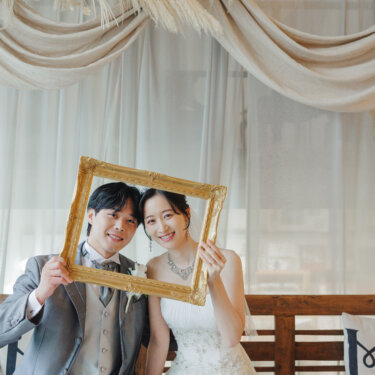 ドレープのセットのベンチで、金の額縁を掲げて笑う新郎新婦。'Mrs'のクッションがかわいいウェディングフォト