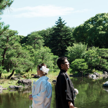 青空と松、池を背に背中合わせで立つ白無垢の花嫁と紋付袴の新郎。白鳥庭園の和装ロケーションフォト