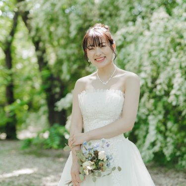 白い花の咲く木立の前でブーケを持ってほほえむ、ビスチェのウェディングドレスの花嫁のソロショットの前撮り