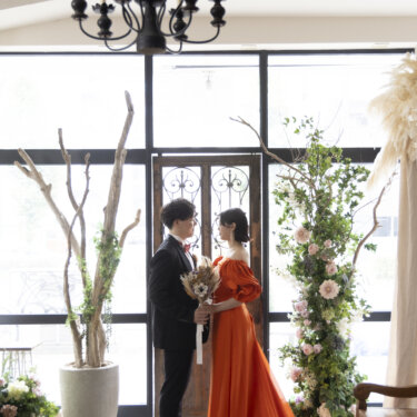 シャンデリアと花のアーチのあるスタジオで向き合う、新郎とトレーンを広げたオレンジのカラードレスの花嫁の前撮り
