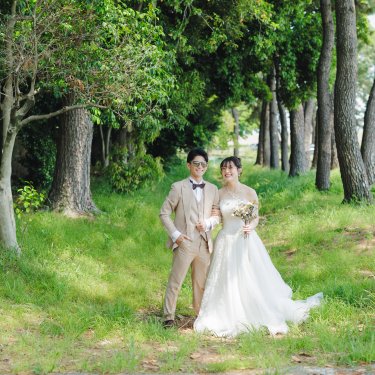 松並木の芝生の小道に並んで立つ、ベージュのタキシードの新郎とウェディングドレスの花嫁を引きで写したロケーションフォト
