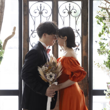 アイアンの窓と花のアーチの前で、額を寄せ合う新郎とオレンジのカラードレスの花嫁のウェディングフォト