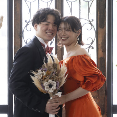 アイアン装飾の窓の前で頬を寄せ、カメラに笑う新郎新婦。ドライフラワーのブーケとオレンジのカラードレスの前撮り