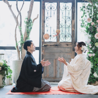 花のアーチと扉のセットで、赤い毛氈に向かい合って座り手毬で遊ぶ新郎新婦。遊び心ある和装前撮り