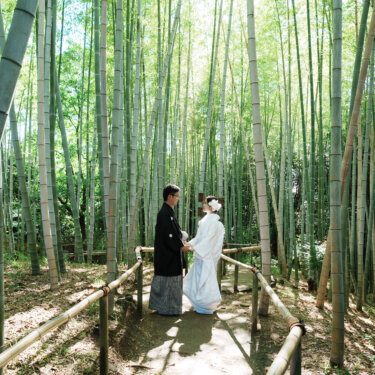高い竹林の中の小道で向き合う紋付袴の新郎と白無垢の花嫁を、引きで写した緑あふれる和装前撮り
