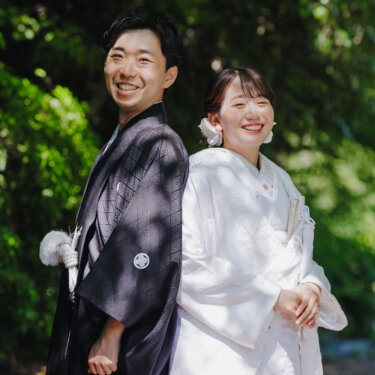 木漏れ日の庭で背中合わせに立ち、カメラに笑顔を向ける紋付袴の新郎と白無垢の花嫁の和装フォトウェディング