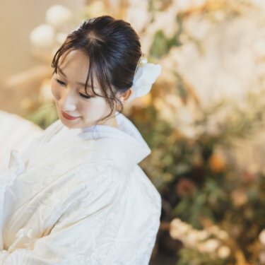 白い和傘を肩にかけ、うつむいてほほえむ白無垢の花嫁のアップ。白い花の髪飾りが映える和装フォトウェディング