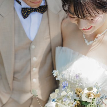 ブーケを見つめてほほえむ花嫁と、寄り添う蝶ネクタイの新郎のアップ。ナチュラルな前撮り