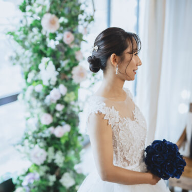 花のアーチのそばで、青いバラのブーケを持って横を向くレースのウェディングドレスの花嫁。シニヨンのヘアアレンジが映える前撮り